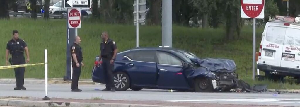 Police officers investigate fatal car accident scene with damaged blue Nissan sedan and white van at Gainesville intersection, yellow tape cordoning area.